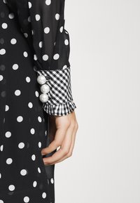 Robe noire à pois avec un tissu texturé et transparent. Elle présente un revers à carreaux avec un bord froufrou et trois grands accents en perles blanches.