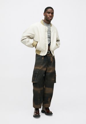 Μπεζ bomber jacket με καφέ γιακά, γκρι μπλουζάκι, cargo παντελόνι με σχέδιο σε μαύρο και καφέ χρώμα, και καφέ μποτάκια με κορδόνια.