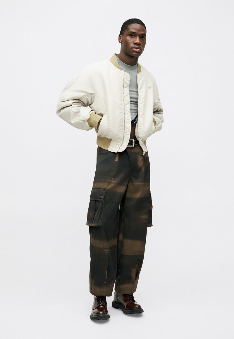 Jachetă bomber bej cu guler maroniu, tricou gri, pantaloni cargo cu model negru și maro, și ghete maro cu șireturi.