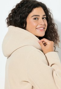 Béžová texturovaná mikina s velkou kapucí, detail límce a ruka upravující látku. Měkký, tkaný materiál.