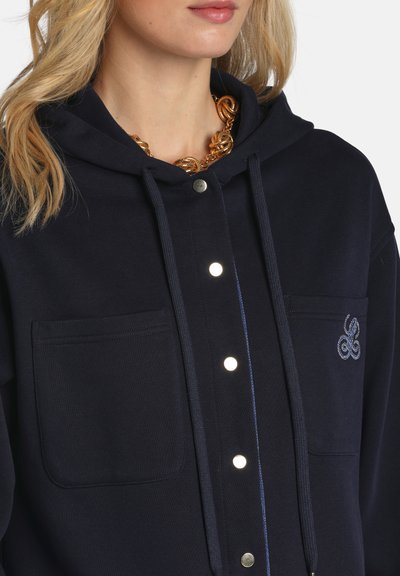 Szőke nő, tengerészkék gombos kapucnis pulóverben, elülső zsebekkel és arany színű, csavart lánc nyaklánccal.