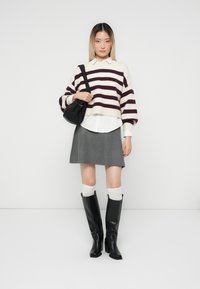 Chunky gebreid sweater met bordeauxstrepen op crème, gelaagd over een witte blouse, gecombineerd met een grijze rok en zwarte kniehoge laarzen.