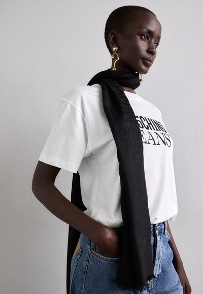 Black scarf with subtle logo detail drapes around the neck, paired with a white t-shirt and high-waisted blue denim jeans.