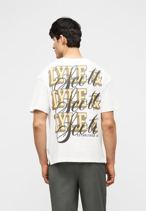 Homme de dos portant un T-shirt blanc avec un grand texte en couches "LYLE & SCOTT" en jaune et noir au dos, pantalon gris, fond uni.