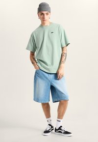 Grøn bomulds t-shirt med et lille logo, sat sammen med lyseblå denim shorts. Bærer en grå strikket beanie og sorte sneakers med hvide detaljer.