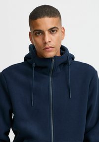 Marineblauwe fleece hoodie met een volledige rits, trekkoordcapuchon en een zachte textuur. Beschikt over een relaxte pasvorm en kangoeroezakken.