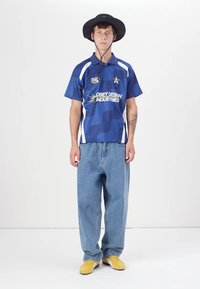 Blue camo-patterned jersey with white accents, worn with loose light blue jeans and yellow sneakers. Hat with chin strap completes the outfit.