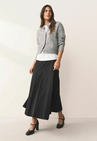 Cardigan gri tricotat, top alb, fustă maxi plisată neagră și pantofi negri cu toc. Texturi netede și linii clare pe tot parcursul ținutei.