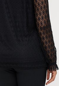 Μαύρη δαντελένια μπλούζα με κυκλικό σχέδιο, μακριά διάφανα μανίκια και σταθερή μαύρη επένδυση από κάτω, με λεπτομέρεια στο τελείωμα σε κόκκινη γραμμή.