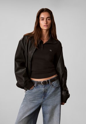 Femme aux longs cheveux bruns portant une veste en cuir noire sur un haut court noir et un jean bleu ample avec une ceinture noire sur un fond gris.