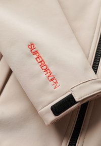 Lichtbeige jasje met een gestructureerde stof, een zwarte rits en een levendig rood geborduurd logo "SUPERDRY/JPN" op de mouw.