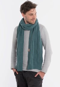 Turkusowy dziany szalik o teksturowanym wzorze, wykonany z miękkiego materiału, zarzucony na szyję, z małą metką marki.