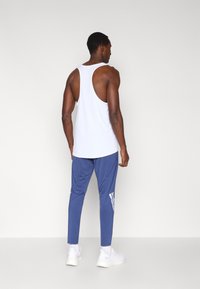 Λευκή αμάνικη μπλούζα με στρογγυλή πλάτη, σε συνδυασμό με μπλε παντελόνι στενής εφαρμογής με λευκές πλευρικές ρίγες και λευκά αθλητικά παπούτσια.