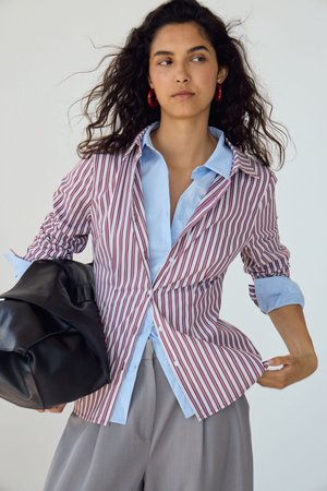 Femme aux cheveux bouclés porte un blazer rayé rouge et blanc sur une chemise bleue, un pantalon gris, des boucles d’oreilles rouges en forme d’anneau, tenant un sac en cuir noir.
