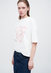 Camiseta blanca de algodón con un gráfico rosa de un león y texto estilizado en el frente. Manga corta y ajuste relajado. Llevada con jeans azules.