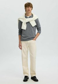 Hallide pikkade varrukatega särk, kreemivärvi kampsun kaela ümber seotud, kreemivärvi püksid ja mustad kingad. Minimalistlik disain pehmete tekstuuridega.