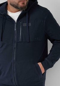 Marineblauwe hoodie met rits aan de voorkant, capuchon en kangoeroezakken. Bevat een gestructureerd paneel en een logo-opdruk op de borst. Zachte, casual stof.