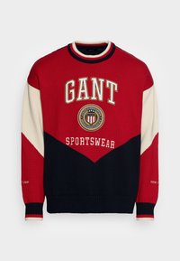 Sweter z dzianiny w kolorach czerwonym, granatowym i kremowym z trójkątnym wzorem. Logo tekstowe "GANT" i "SPORTSWEAR" na środku oraz na detalu rękawa.