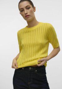 Geel gebreid sweater met korte mouwen met een gestructureerd patroon en geribde halslijn. Gepaarde met donkere denimjeans.