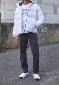 Bílá péřová bunda s kapucí, zapínání na zip; v kombinaci s bílým mikinou s logem, šedými džíny a bílými sportovními botami.
