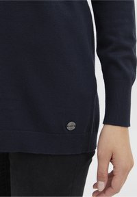 Mörkblå stickad tröja med långa ärmar, ribbade muddar och fåll; har en cirkulär detalj i grått metalllogo på sidan.