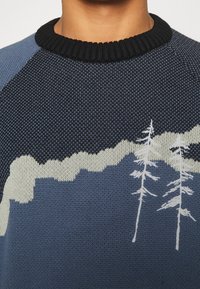 Gros plan d'un pull en tricot bleu avec un col noir présentant des motifs blancs d'arbres et de montagnes sur la poitrine.