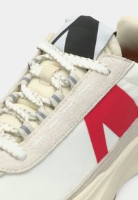 Αθλητικό παπούτσι με μπεζ επάνω μέρος από σουέντ, λευκό υφασμάτινο σώμα, κόκκινο τρίγωνο λογότυπο, γκρι λεπτομέρειες και κορδόνια με υφή. Μαύρη και λευκή θηλιά στην πτέρνα.