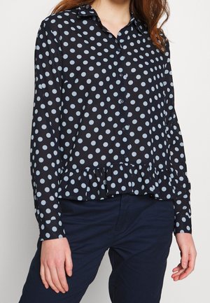 Kobieta ubrana w granatową koszulę z długim rękawem, zapinaną na guziki, z białymi kropkami i falbanką na dole, w połączeniu z ciemnogranatowymi spodniami.