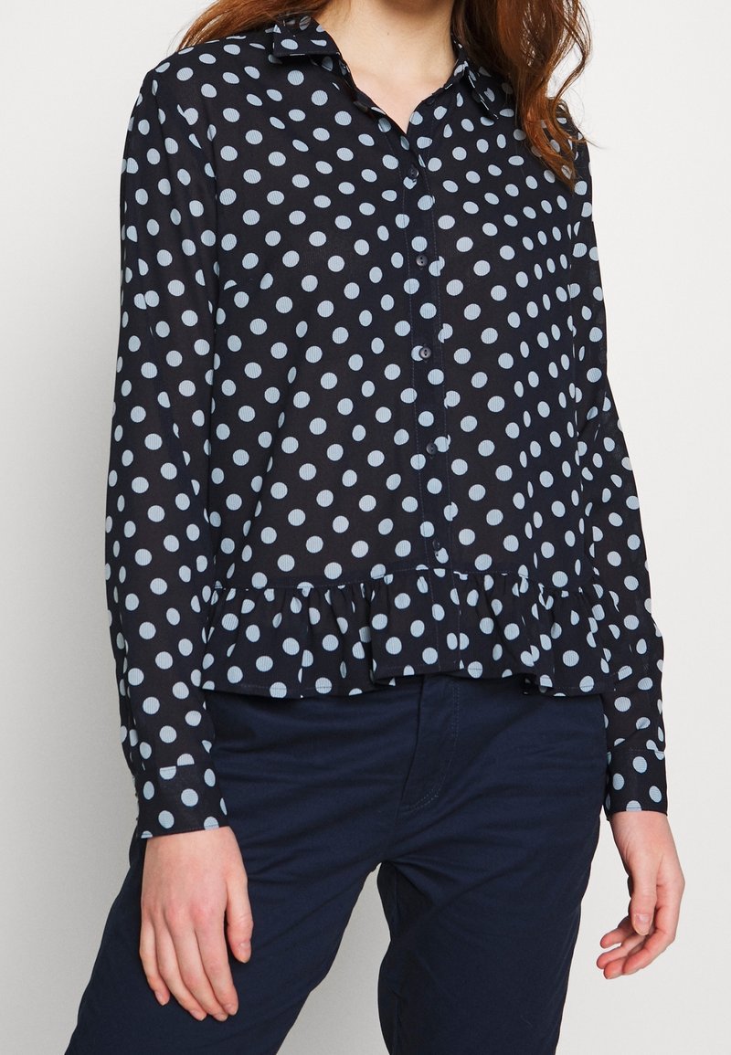 Femme portant un chemisier à manches longues bleu marine à pois blancs avec un ourlet à volants, associé à un pantalon bleu marine foncé.