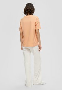 Pfirsichfarbene Baumwoll-T-Shirt mit kurzen Ärmeln, lockerer Passform und geradem Saum, kombiniert mit weißen Culottes und weißen Schuhen.