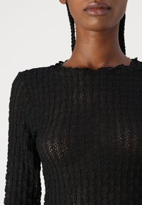 Black textured top with long sleeves, featuring detailed lace-like pattern and scalloped neckline. Material appears lightweight and semi-transparent.