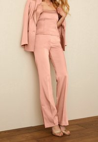 Costume rose pâle composé d'un blazer ajusté, d'un top sans manches et d'un pantalon droit en tissu lisse. Des sandales dorées à bout ouvert complètent le look.
