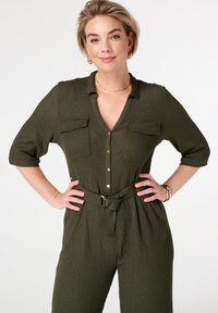 Combinaison en vert olive avec un tissu texturé, fermeture à boutons sur le devant, deux poches poitrine, manches courtes et taille ceinturée avec une boucle circulaire.