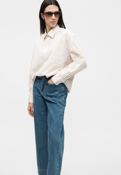 Γυναίκα που φοράει μπεζ ριγέ πουκάμισο με κουμπιά, μπλε τζιν με ψηλή μέση και φαρδιά μπατζάκια με μεγάλες μπροστινές τσέπες και καφέ μεγάλα γυαλιά ηλίου.