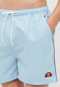 Bañador azul claro hecho de material sintético, con cintura elástica, cierre de cordón, bolsillos laterales y un parche de logo en contraste.