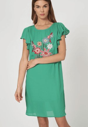 Mujer con vestido verde de manga corta con mangas con volantes y flores bordadas en rojo, blanco y azul con pájaros en el pecho.