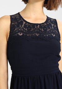 Marinblå klänning med en genomskinlig spetsöverdel som har blommig broderi. Midjan är åtdragen med ett veckat band, vilket leder till en flödande kjol.