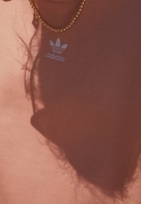 Nära bild av en person som bär en brun Adidas-tröja och ett guldhalsband som kastar en skugga på tyget.