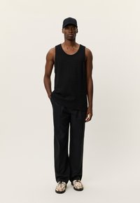 Μαύρο τοπ με υφή, σε συνδυασμό με άνετο μαύρο παντελόνι και παπούτσια με σχέδιο. Το μοντέλο φοράει μαύρο καπέλο, στέκεται μπροστά σε ουδέτερο φόντο.