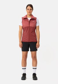 Gilet rosso con zipper frontale e colletto alto, abbinato a pantaloni shorts neri e calze bianche a righe. Scarpe nere completano il look.