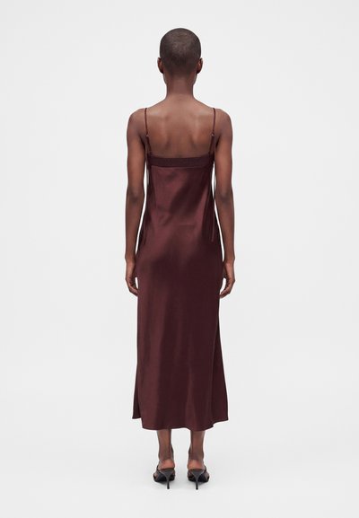 Femme aux cheveux courts portant une robe sans manches en satin bordeaux foncé et des chaussures à talons noirs, vue de dos sur un fond uni.