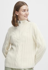 Pull en tricot couleur crème avec un col montant zippé, texture côtelée et accents en point de câble sur le devant et les manches.