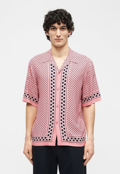 Άνδρας που φοράει ροζ πουκάμισο με κοντά μανίκια και μαύρο σχέδιο καρδιάς, στέκεται μπροστά σε απλό ανοιχτό φόντο.