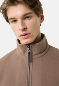 Bruine fleecejas met een hoge kraag en een volledige ritssluiting. Voorzien van een metalen ritstrekker met een merklabel en een zachte textuur.