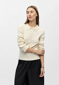 Ribstrikket cremefarvet sweater med bred krave og seks lyseblå knapper. Stylet med sorte bukser med vide ben for kontrast.