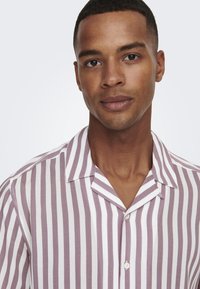 Camisa de manga corta con cuello y rayas verticales en púrpura y blanco. Está confeccionada con una tela ligera y cuenta con un solo botón en el cuello.