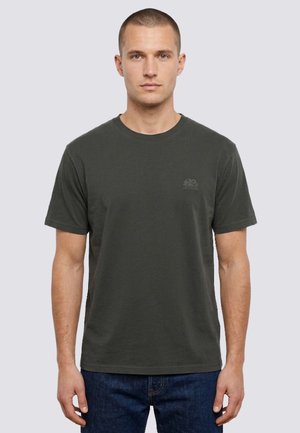 Uomo che indossa una t-shirt semplice grigio scuro a maniche corte e jeans blu, in piedi davanti a uno sfondo grigio chiaro.