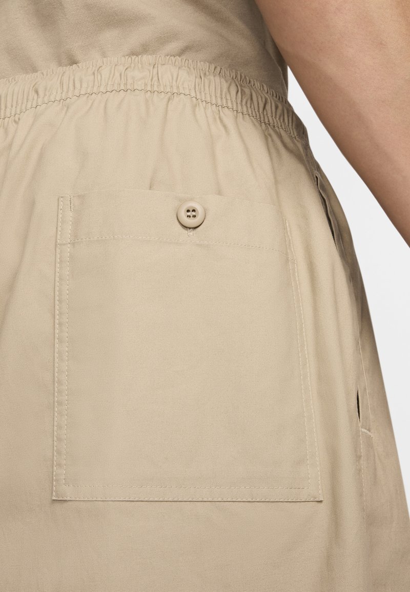 Pantalones cortos de tela beige con cintura elástica y un gran bolsillo cuadrado con detalle de botón en la parte posterior. Textura suave, diseño casual.