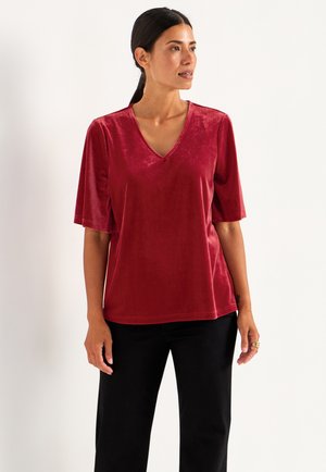 Kurzes rotes Samtoberteil mit V-Ausschnitt, entspanntem Schnitt und glatter Textur. Kombiniert mit schwarzen Hosen, die ein einfaches Design zeigen.