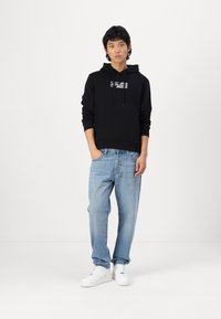 Sudadera con capucha negra y un gráfico blanco. Combinada con jeans azul claro y zapatillas blancas, de pie contra un fondo liso.